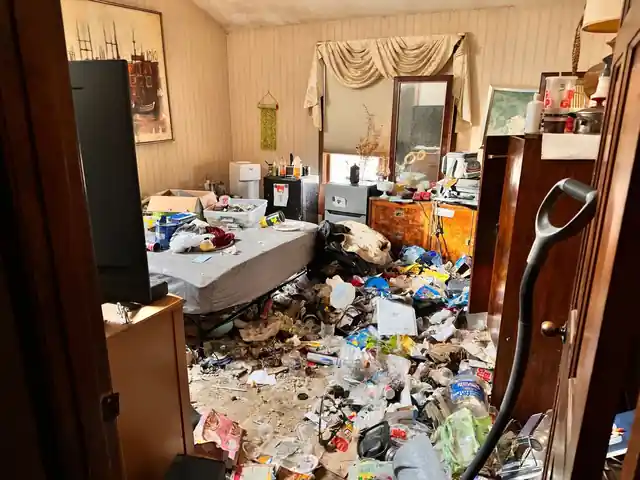 Bedroom with a bare mattress, a wooden dresser and a floor covered in bags, cups and packaging