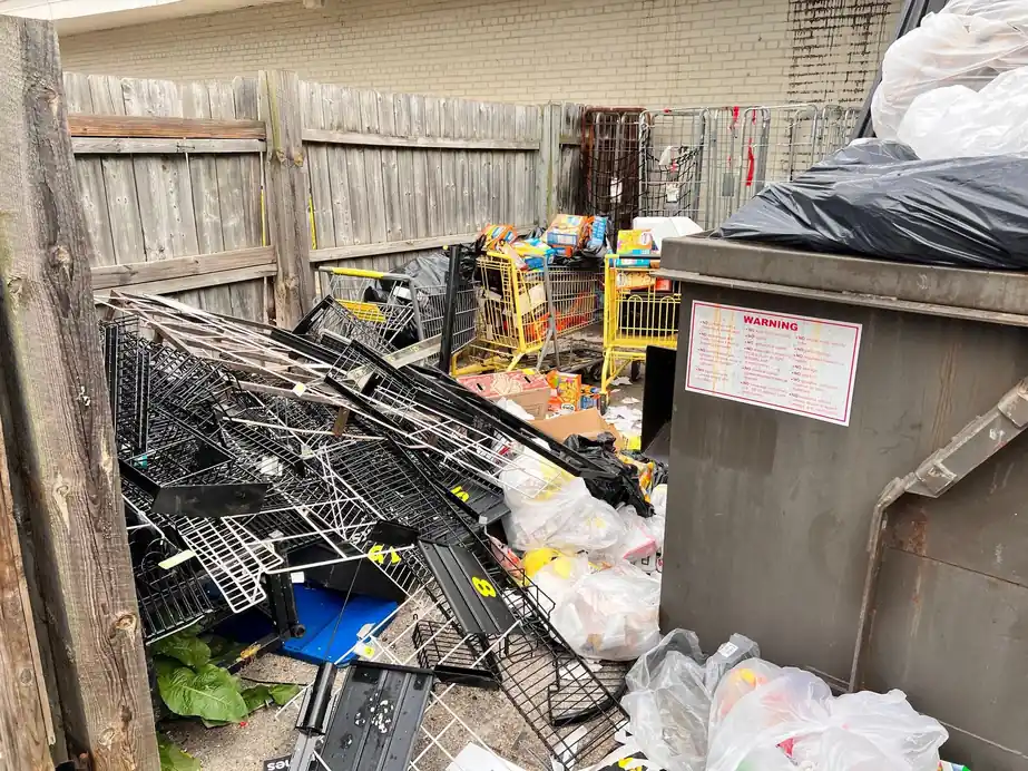 Grey dumpster beside a fence line with wire shelving and bagged debris