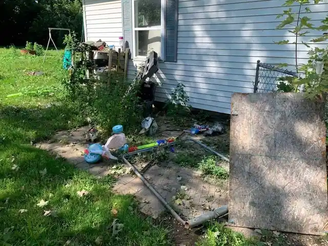 Side yard along white siding with an OSB panel, bent metal pipe and children's toys on the ground