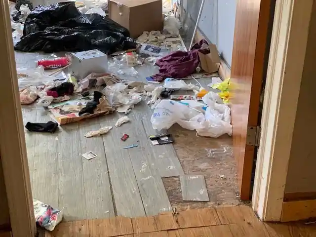 Grey plank floor strewn with plastic bags, clothes and trash by a doorway