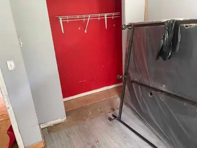 Room with a red wall and a wire shelf above a grey plank floor, a bed frame and mattress standing at the right