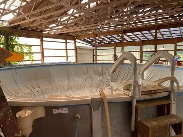 Above-ground pool with a white ladder inside a pole barn with timber framing and clear roof panels