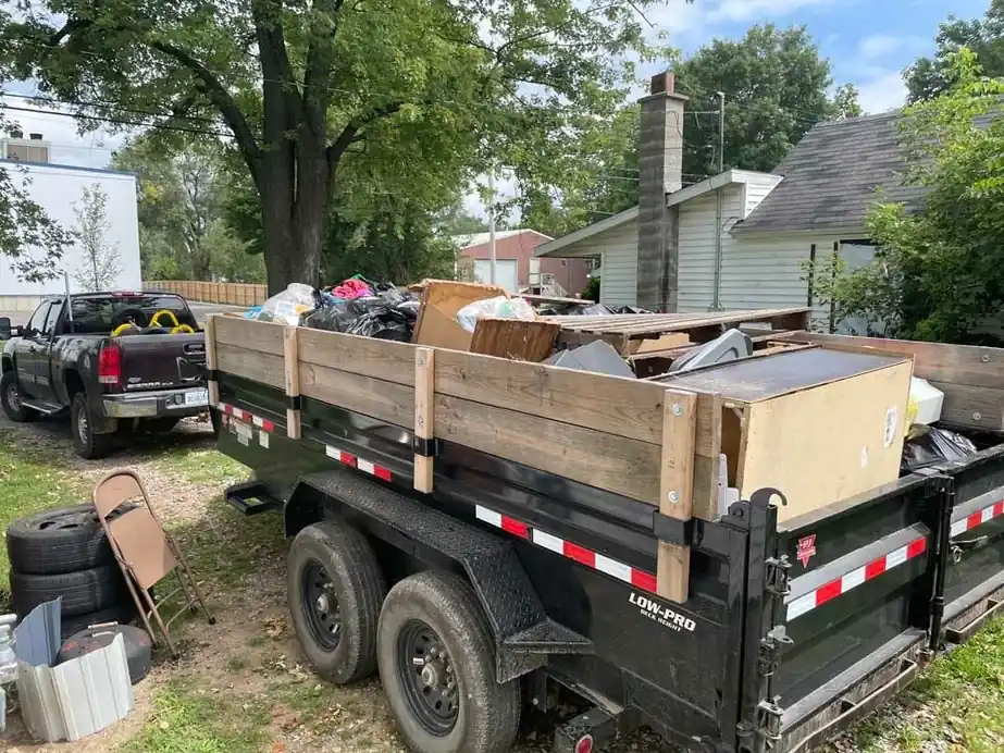 Black dump trailer stacked above its rails with furniture and boxes, pickup behind